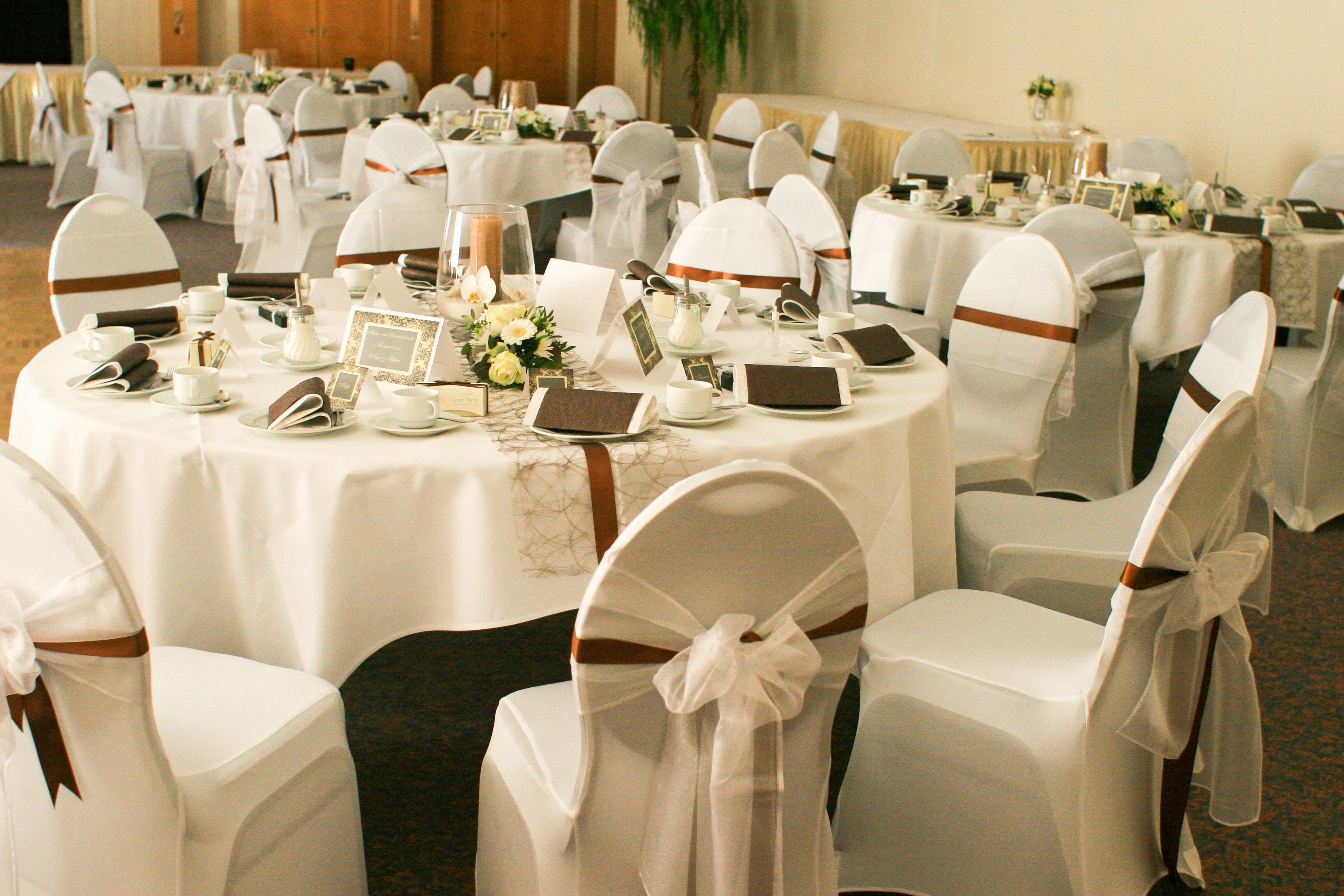 A banquet hall with round tables set for a formal event, each with white tablecloths, chairs with white covers and brown sashes, and table settings including plates, glasses, and cutlery.