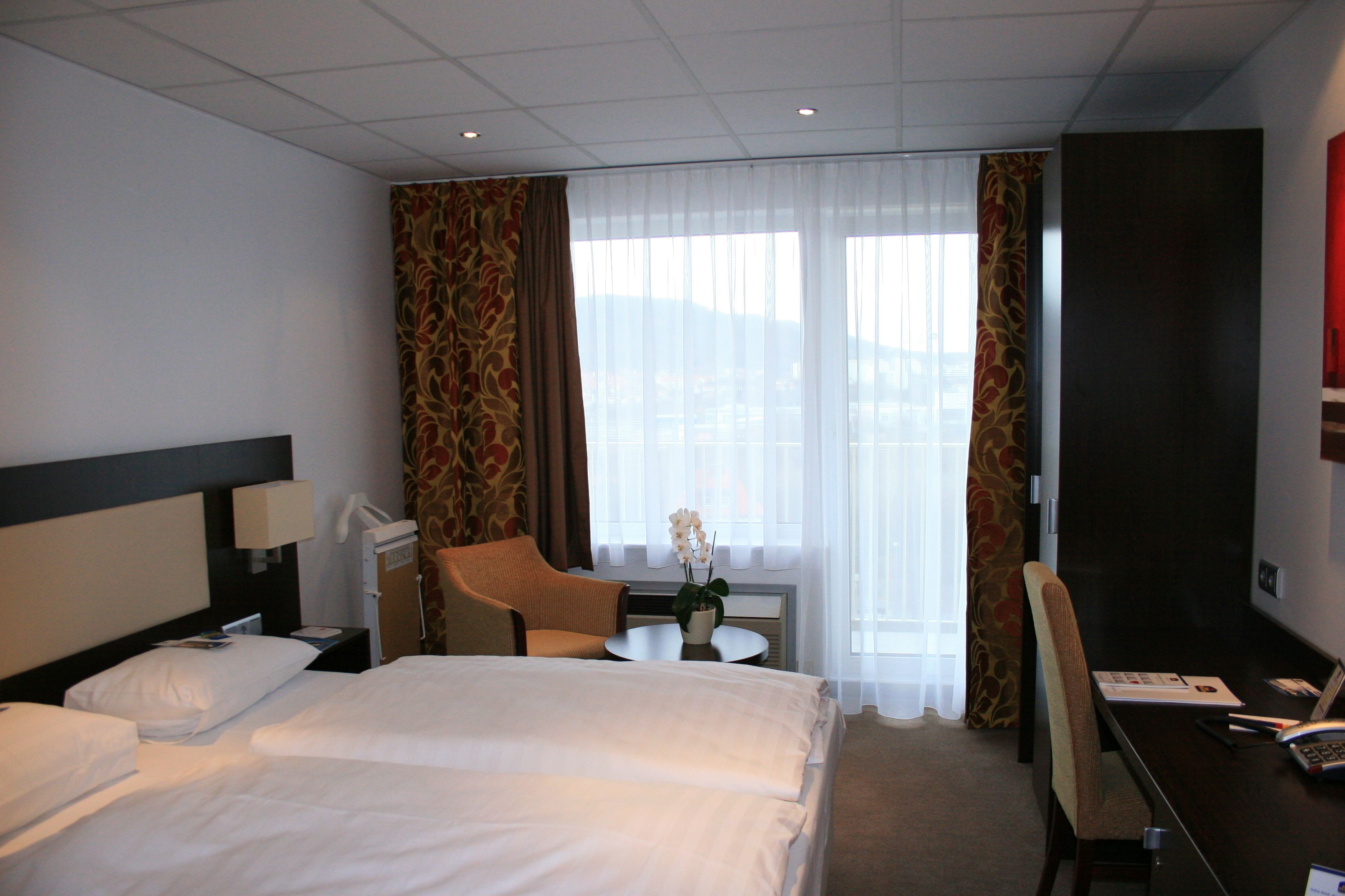 Ein Hotelzimmer mit zwei Betten, einem Schreibtisch, einem Stuhl, einem kleinen Tisch und einem Fenster mit Vorhängen.