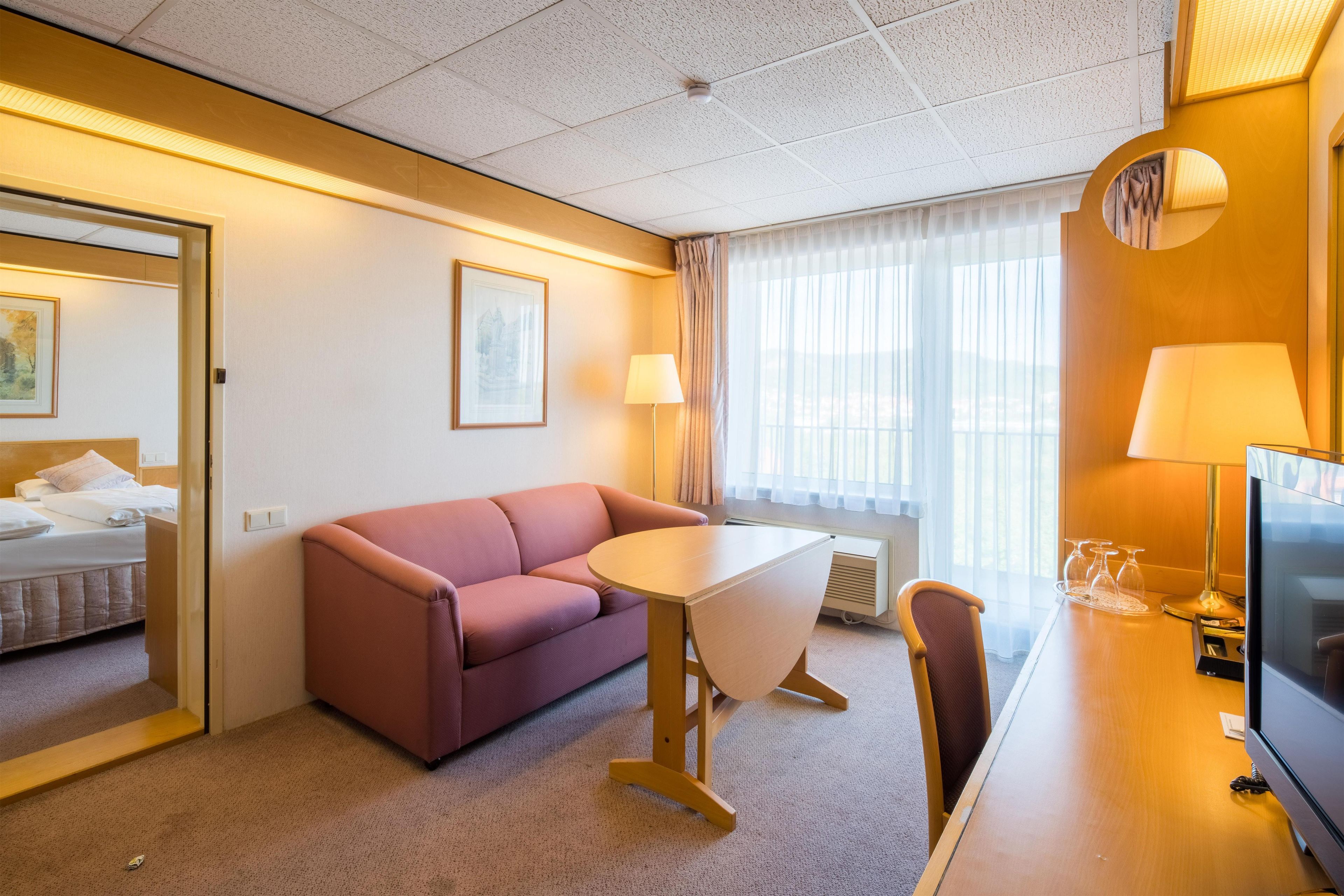 A hotel room with a bed, a sofa, a small table, a chair, a TV, and a window with curtains.