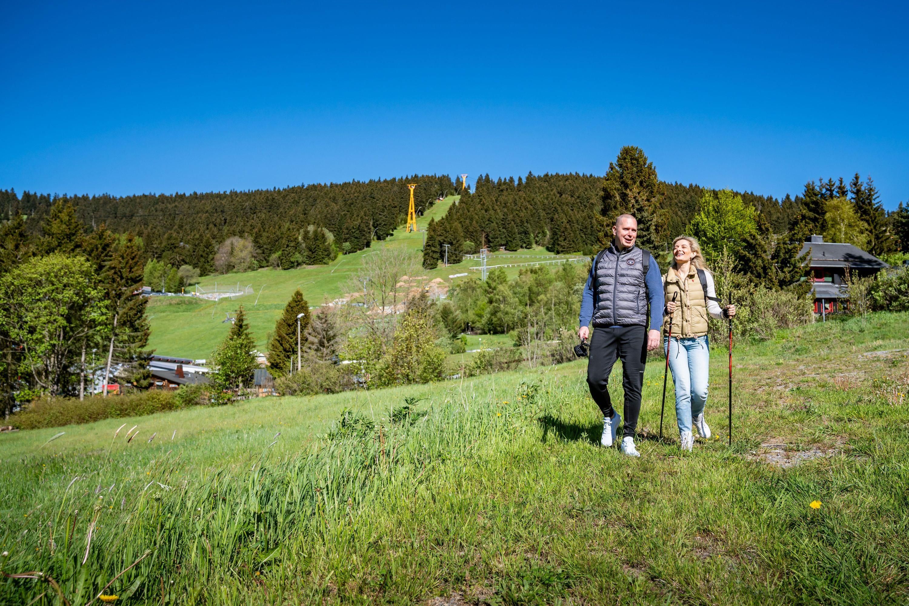 Una pareja está de pie en una colina cubierta de hierba, con un bosque al fondo y un pequeño edificio a lo lejos.
