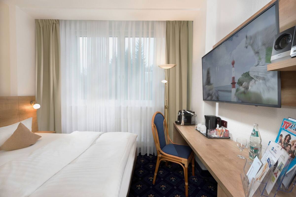 Une chambre d’hôtel comprenant un lit, un bureau avec une chaise, une télévision et une fenêtre avec des rideaux.