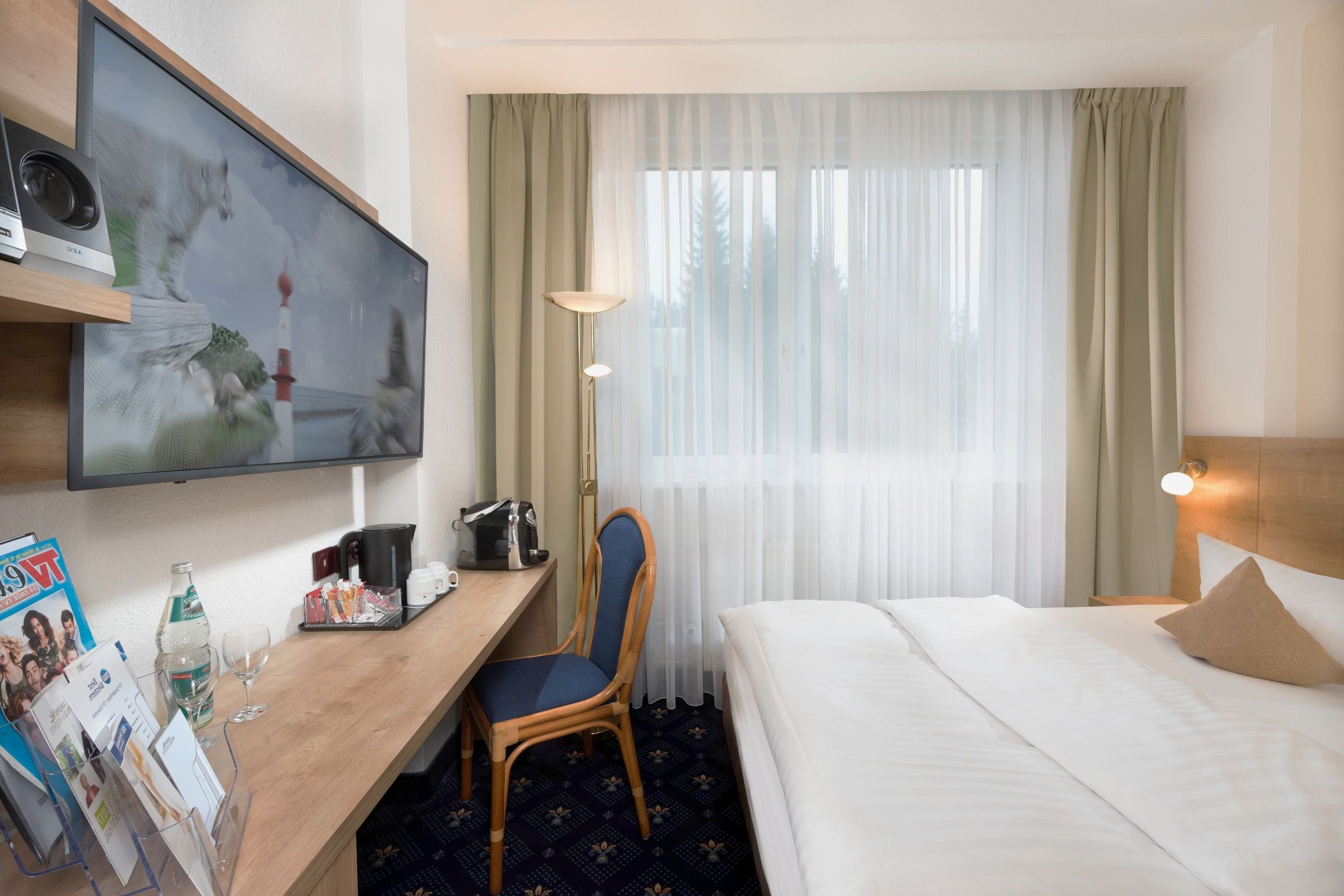 Ein Hotelzimmer mit Bett, Schreibtisch mit Stuhl, Fernseher, Lampe und einem fenster mit Vorhängen.
