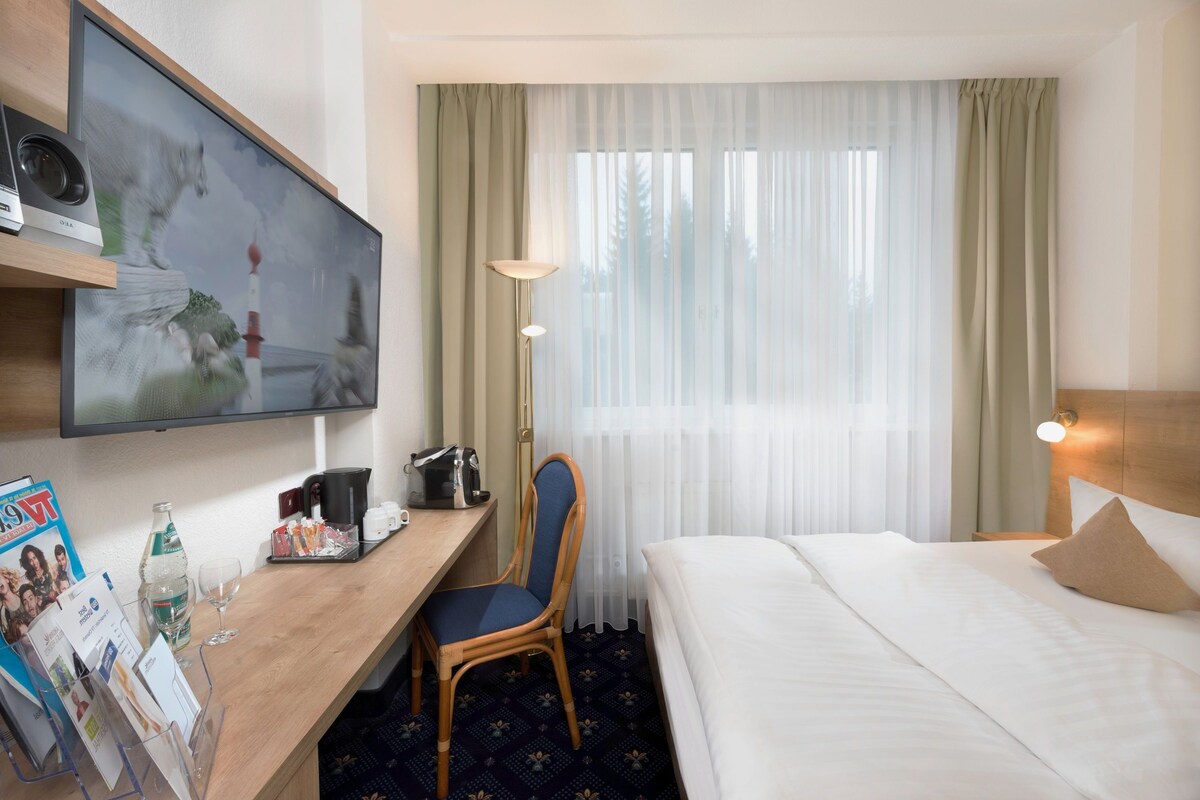 A hotel room with a bed, a desk with a chair, a television, a lamp, and a window with curtains.
