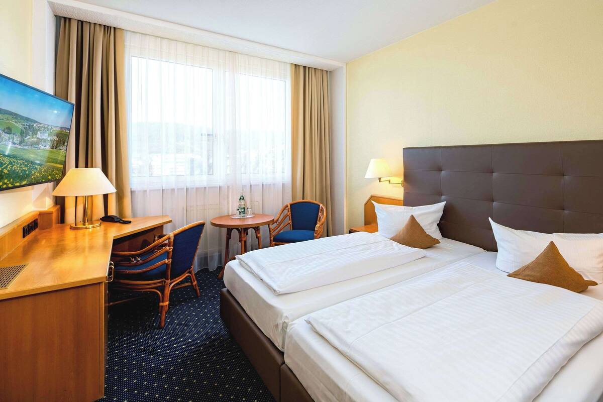 Une chambre d’hôtel avec deux lits, un bureau, une chaise, une télévision et une fenêtre avec des rideaux.
