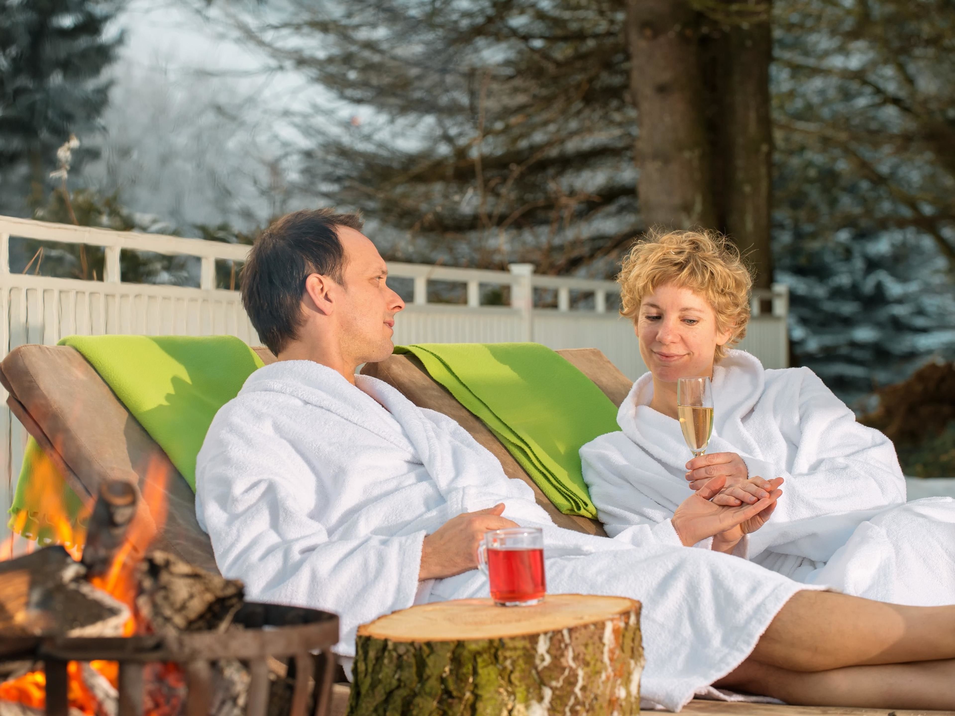 Two people in white robes sitting on lounge chairs by a fire, with a wooden stump and a glass of drink on a table.
