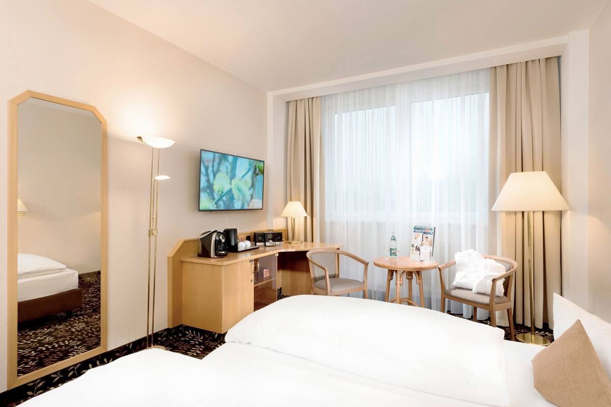 A hotel room with a bed, a desk with a coffee maker, a chair, a table with a magazine, and a large window with curtains.