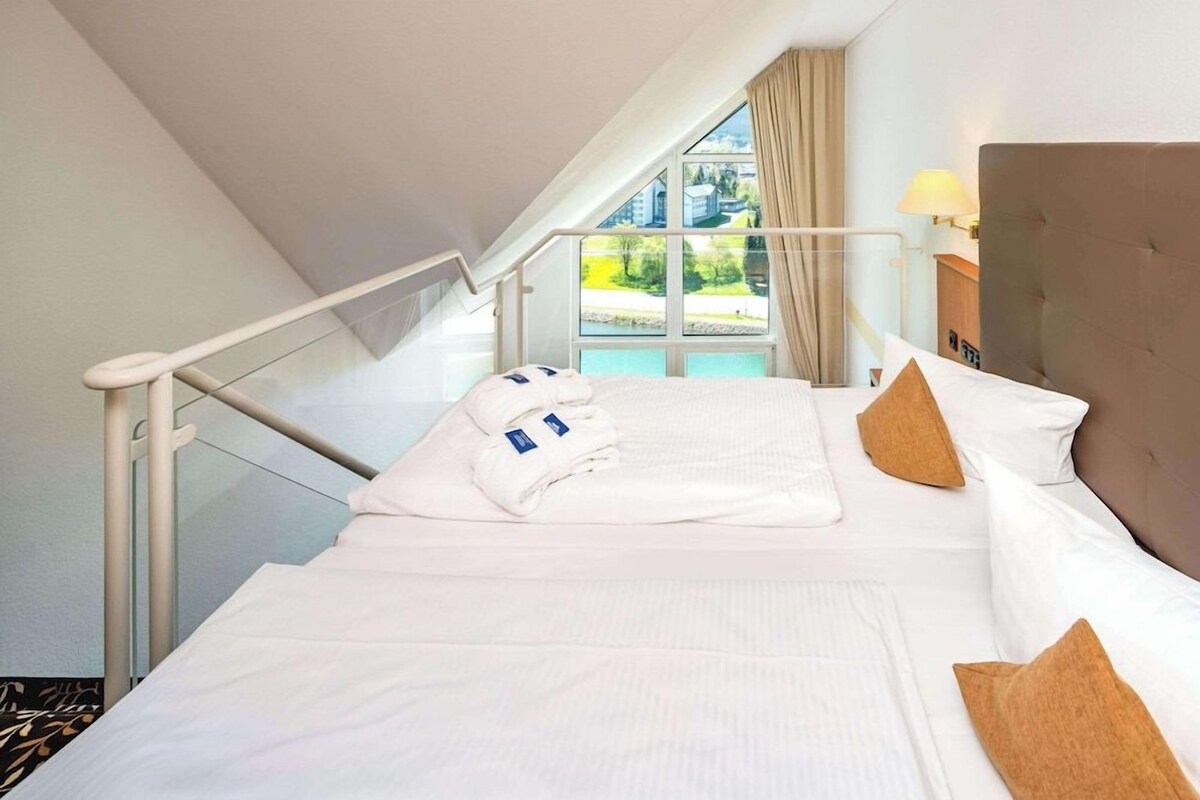 A hotel room with a bed, a window with a view, a staircase, and a lamp.