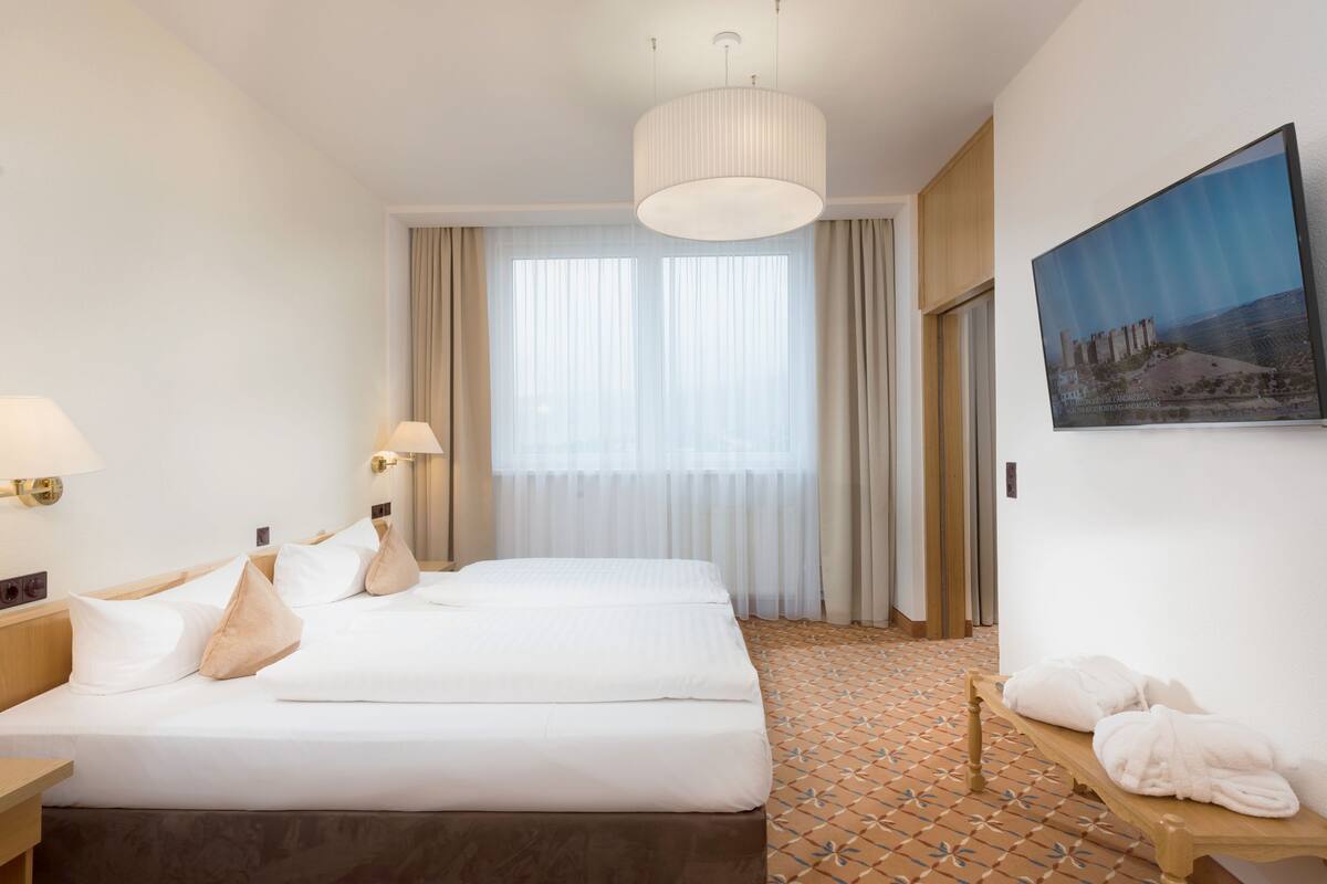 Une chambre d’hôtel avec deux lits, une grande fenêtre avec des rideaux, un tableau au mur et un sol carrelé.