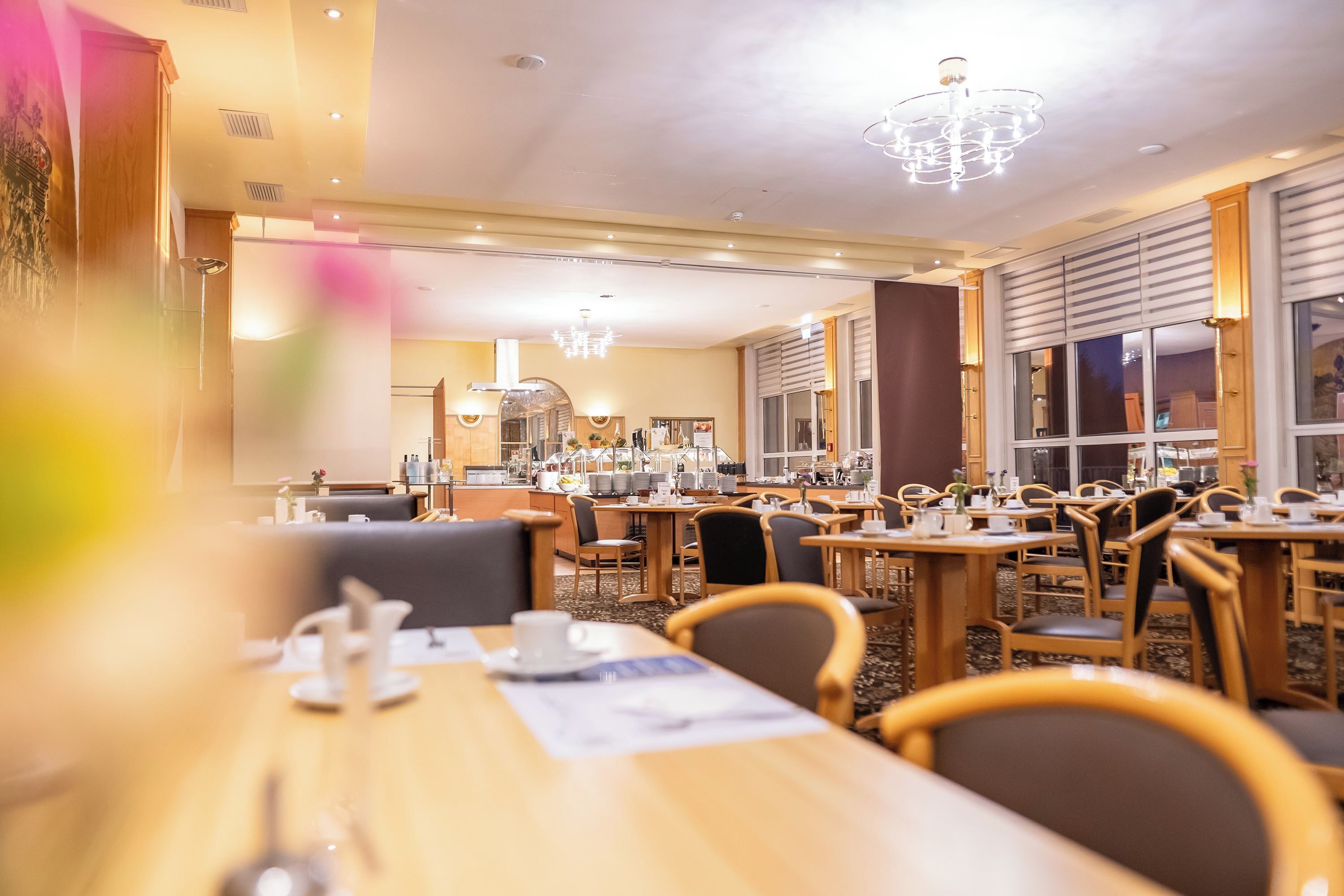 Ein geräumiger Hotel-Speisesaal mit mehreren Tischen und Stühlen, großen Fenstern und einer Kronleuchter.