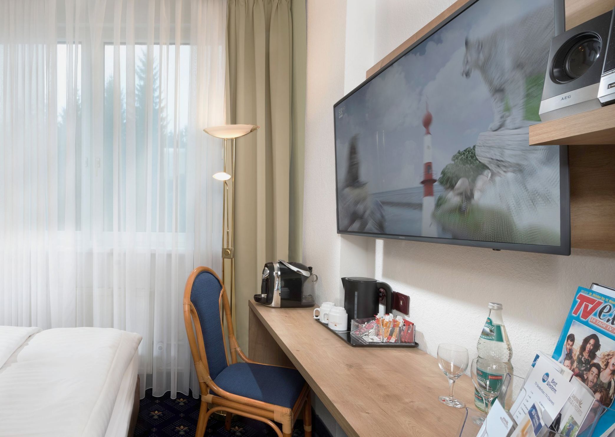 Habitación de hotel con cama, escritorio con cafetera, televisión y un mueble para revistas.