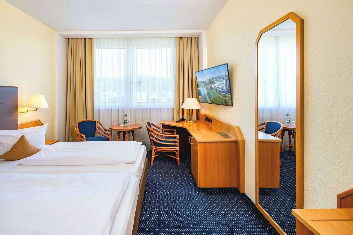 Une chambre d’hôtel avec deux lits, un bureau, une chaise, une petite table, un miroir et une fenêtre avec des rideaux.