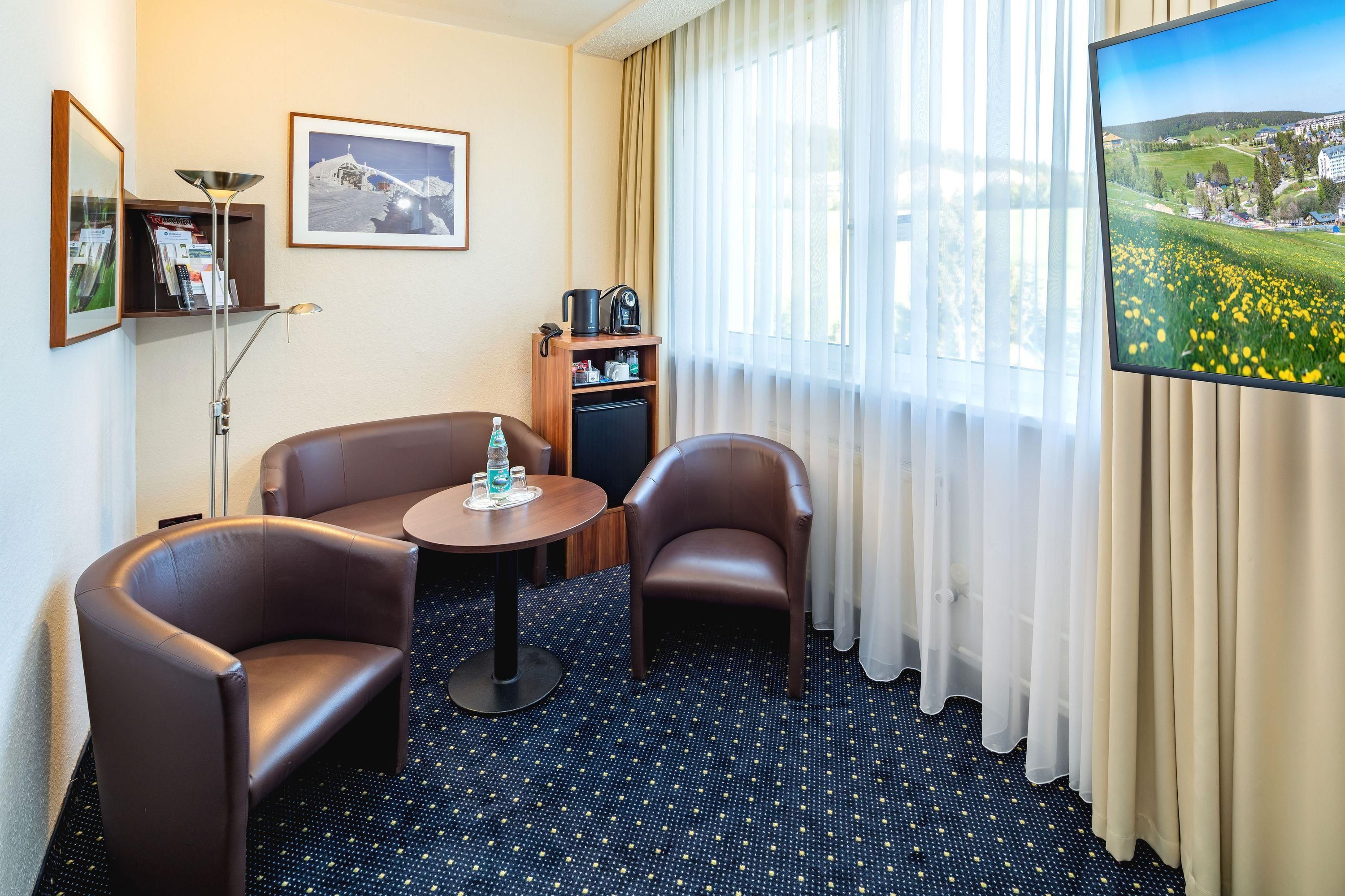 A hotel room with two leather armchairs, a small round table, a minibar, a painting, and a large window with a view of a green landscape.