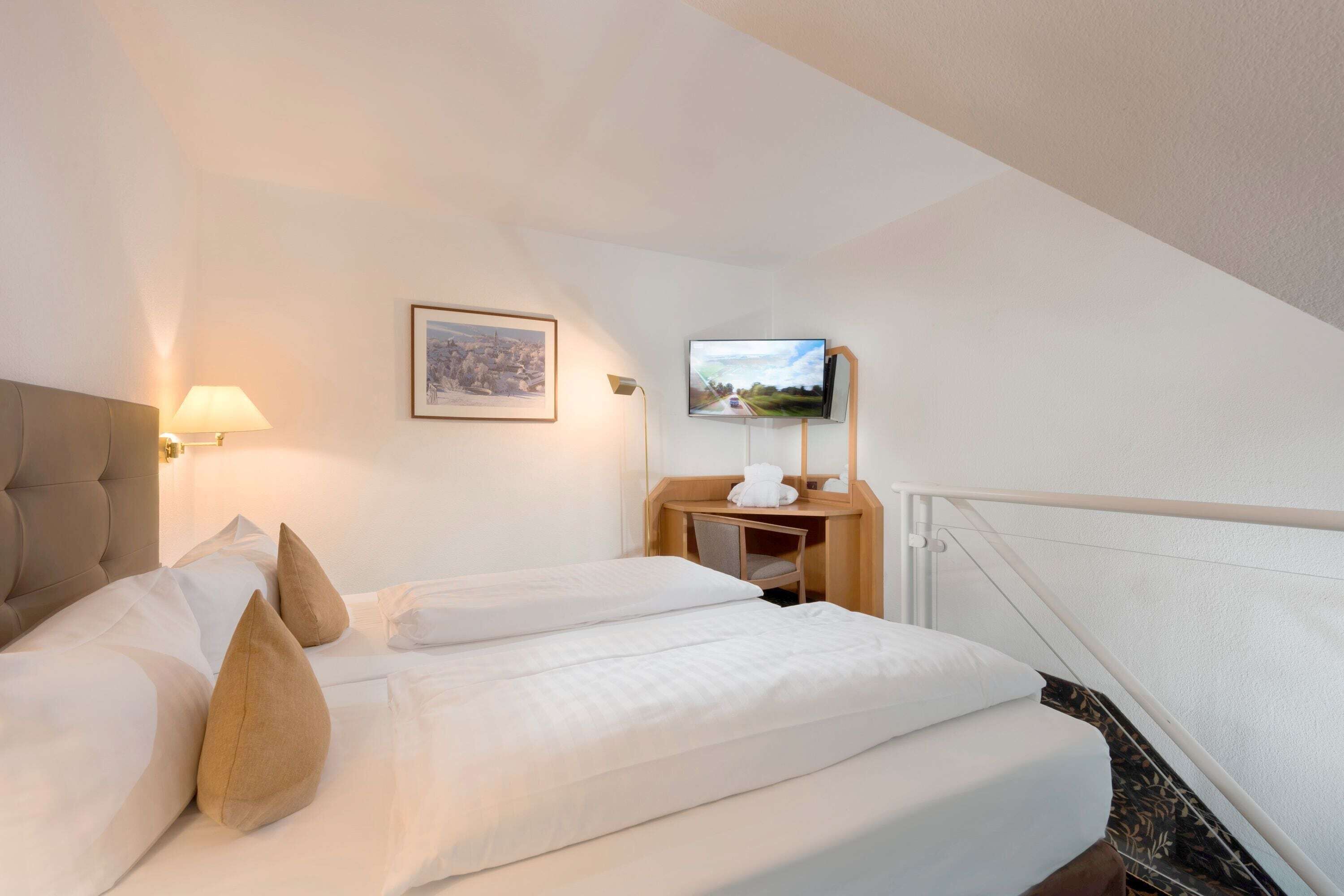 A hotel room with a bed, a desk, a chair, a lamp, a framed picture, and a TV.