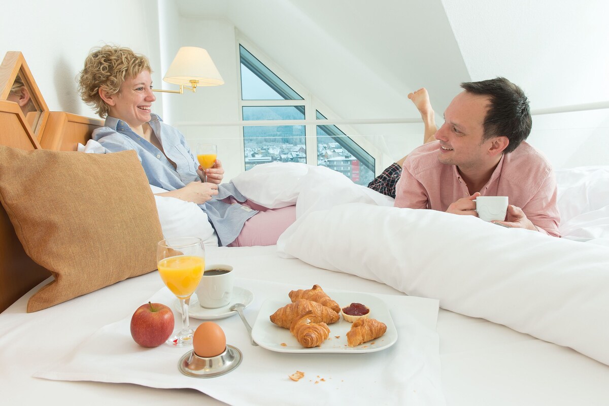 A couple in a bedroom with a breakfast setup including croissants, an apple, orange juice, and coffee.