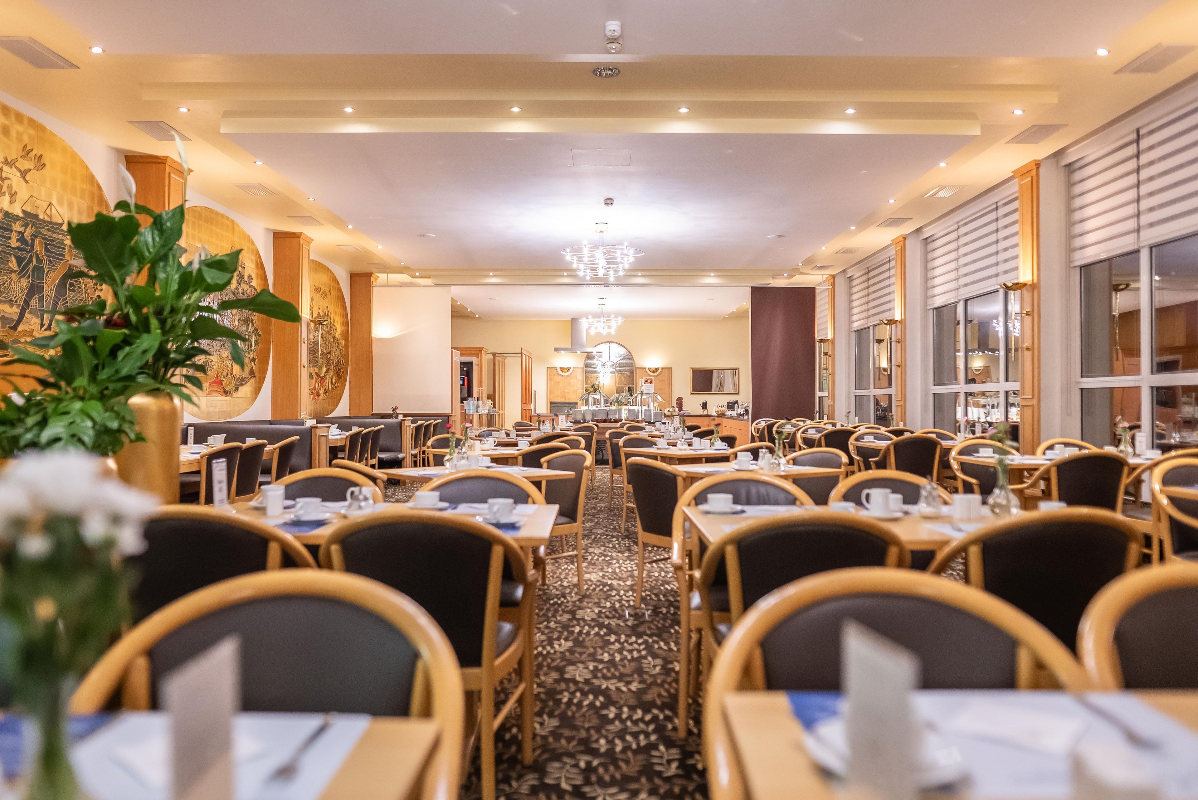 En restaurant i et hotel med flere borde og stole, en stor lysekrone i midten og et mønstret tæppe.