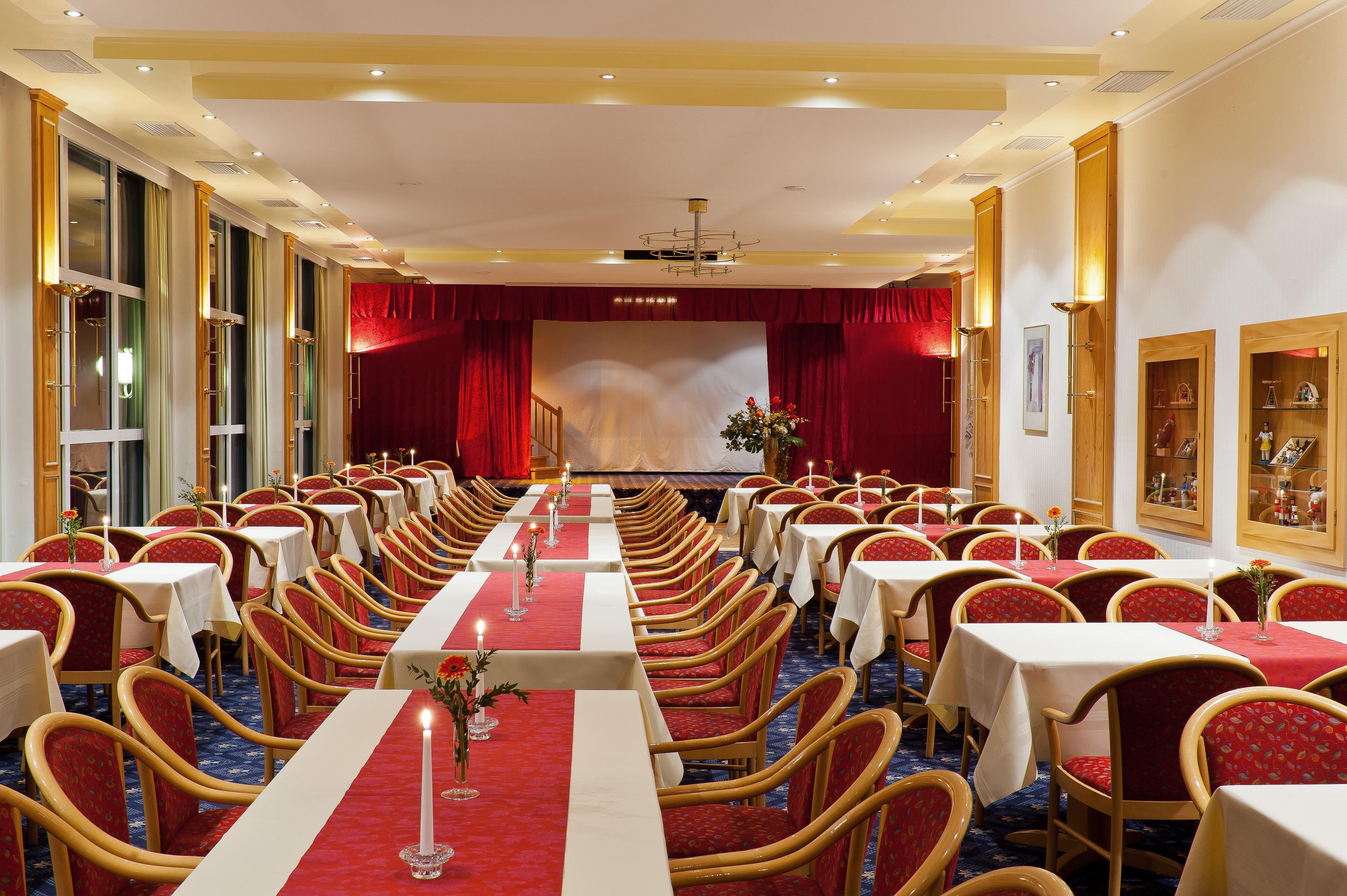 A conference room with rows of tables and chairs, a stage with red curtains, and a chandelier.