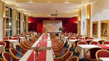 A conference room with rows of tables and chairs, a stage with red curtains, and a chandelier.