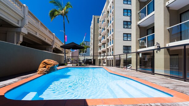 A swimming pool with a wooden deck, a palm tree, and a building with balconies. - Riverside Hotel Southbank (South Brisbane)