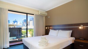 A hotel room with a large bed, a nightstand, a dresser, and a view of a cityscape through a large window. - Riverside Hotel Southbank (South Brisbane)
