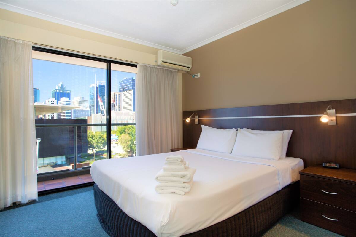 Une chambre d’hôtel avec un grand lit, une table de chevet, une commode et une vue sur le paysage urbain, visible à travers une grande fenêtre.