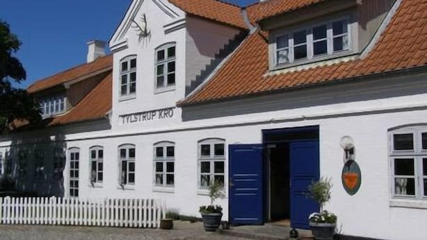 Un bâtiment blanc avec un toit recouvert de tuiles rouges et une porte bleue, portant l’inscription « TYLSTRUP KRO ».