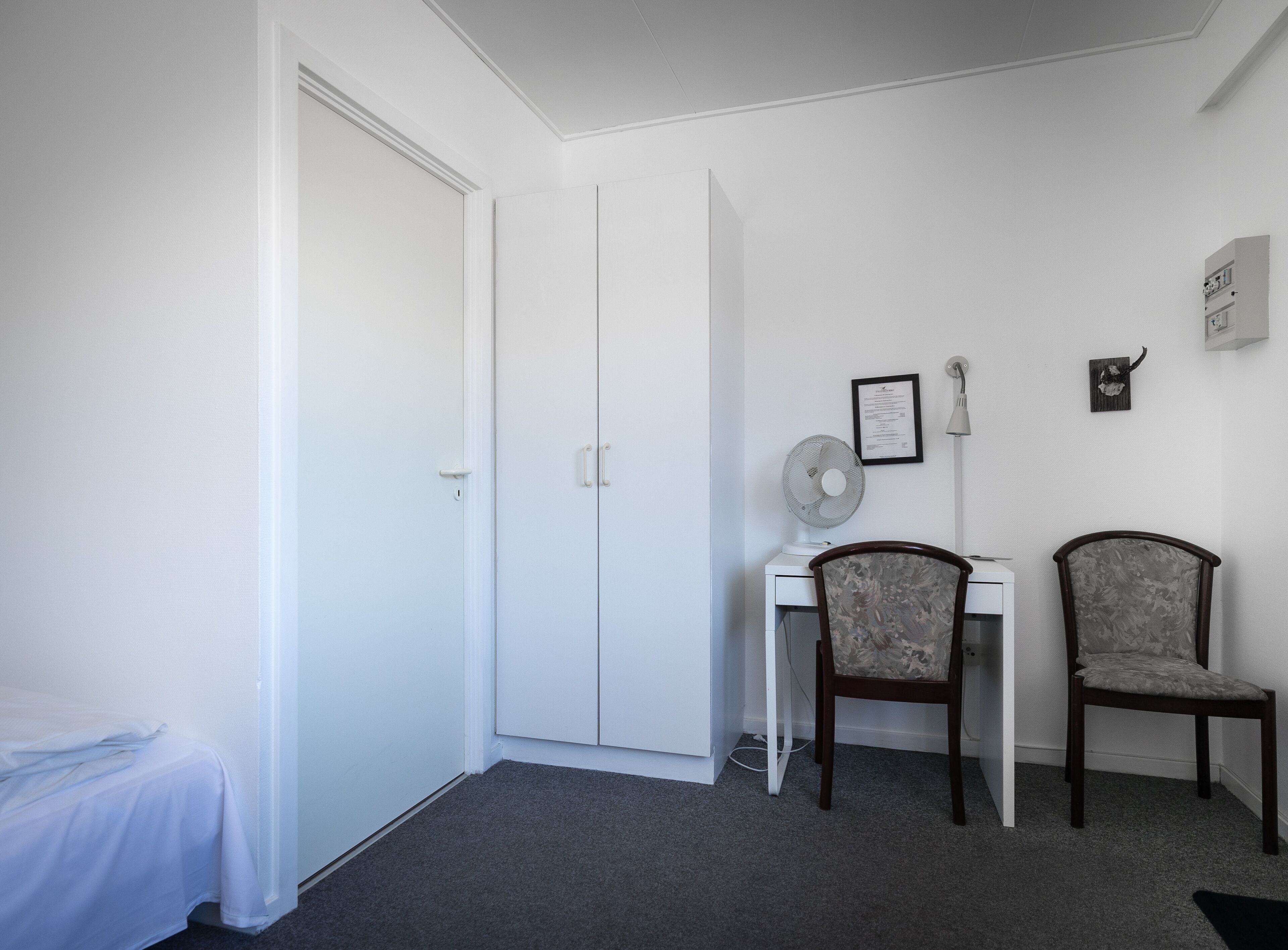 A small, clean room with a bed, a desk, two chairs, a standing fan, and a white wardrobe.