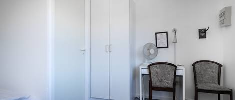 Una habitación pequeña y limpia con cama, escritorio, dos sillas, un ventilador y un armario blanco.