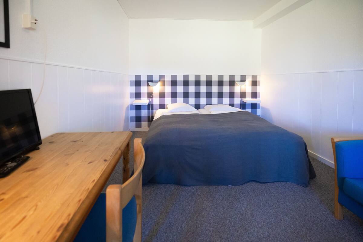 Une petite pièce, bien rangée, avec un lit, un bureau et une chaise. Il y a une télévision sur le bureau et les murs sont blancs.