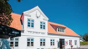 Un bâtiment blanc avec un toit rouge, et l’inscription « TYLSTRUP KRO » sur la façade. On y trouve des tables et des chaises à l’extérieur, devant l’entrée, ainsi qu’un chemin de gravier menant à l’entrée.