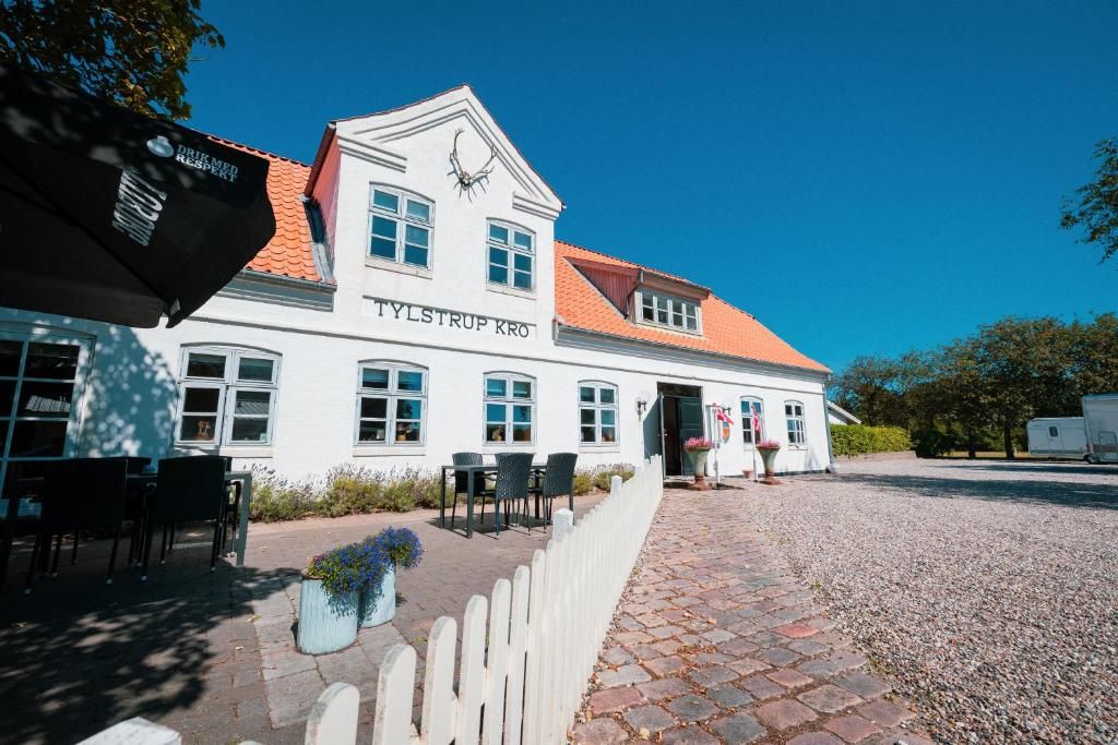Ein weißes Gebäude mit der Aufschrift 'TYLSTRUP KRO' an der Fassade, das ein Ziegeldach, einen gepflasterten Eingangsbereich mit Außenplätzen und einen Schotterweg hat.