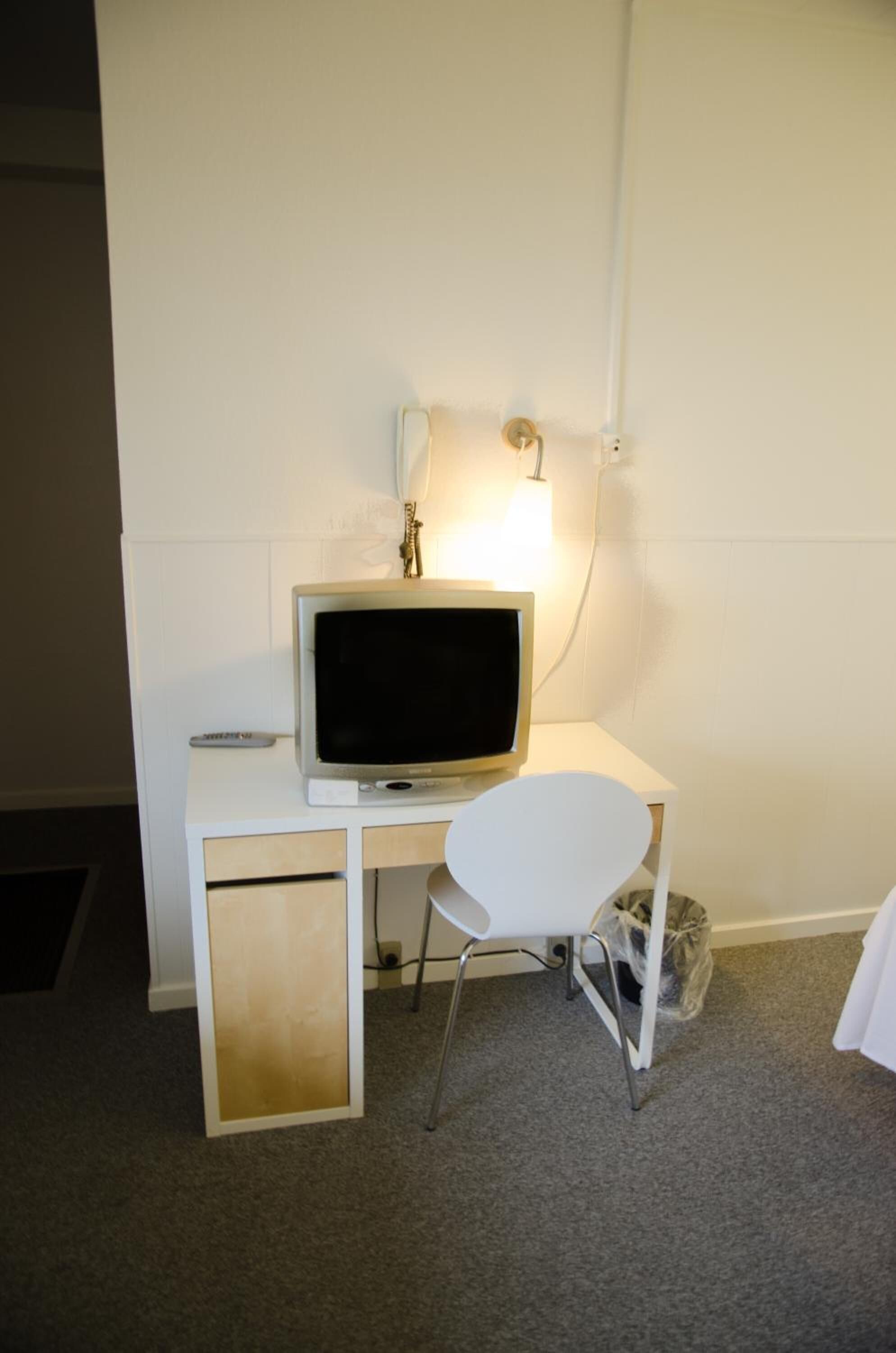 Uma pequena mesa branca com uma televisão, uma cadeira e uma luminária fixada na parede.