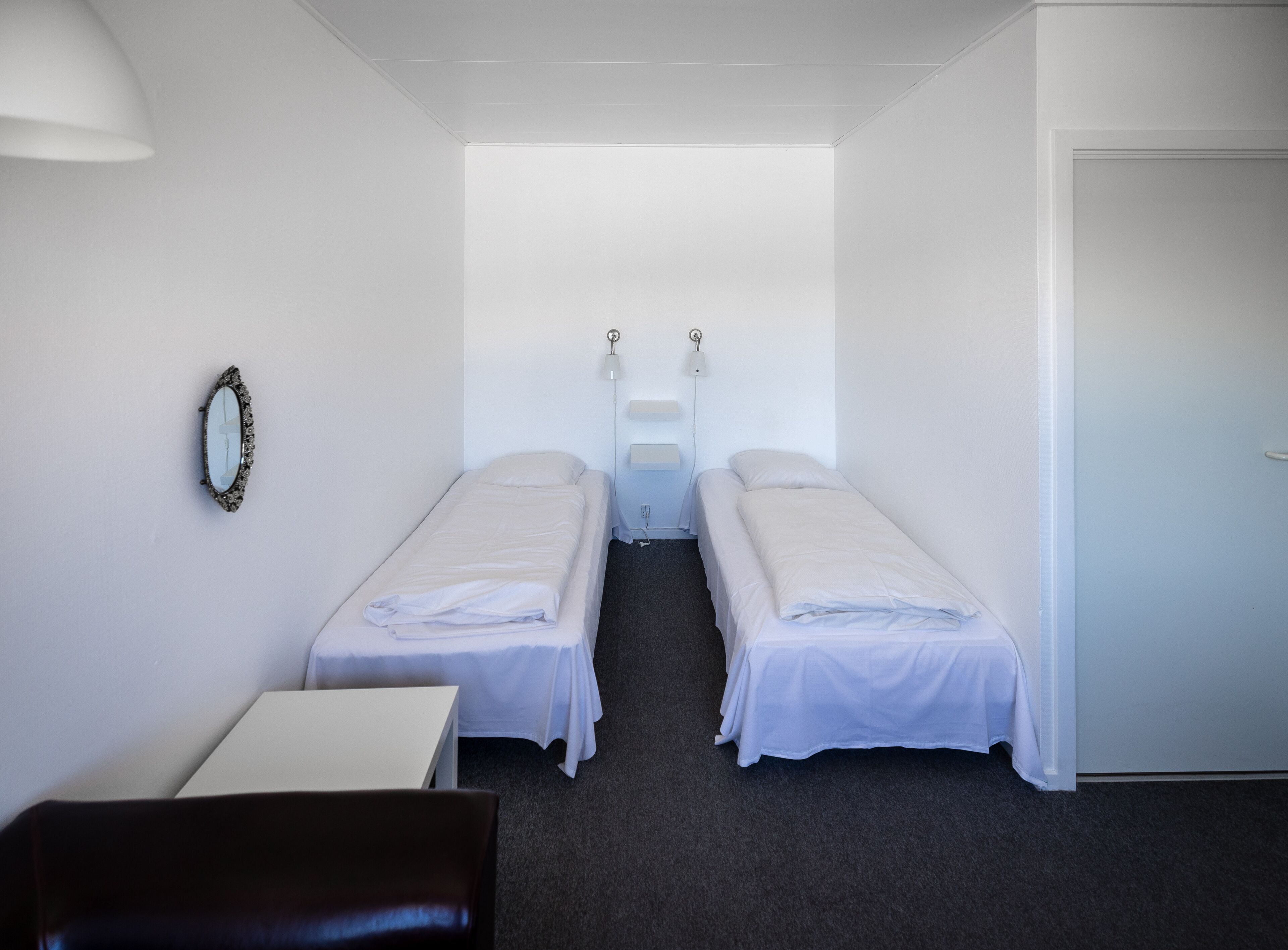 Una habitación pequeña y limpia con dos camas individuales, un escritorio, una silla y un espejo en la pared.