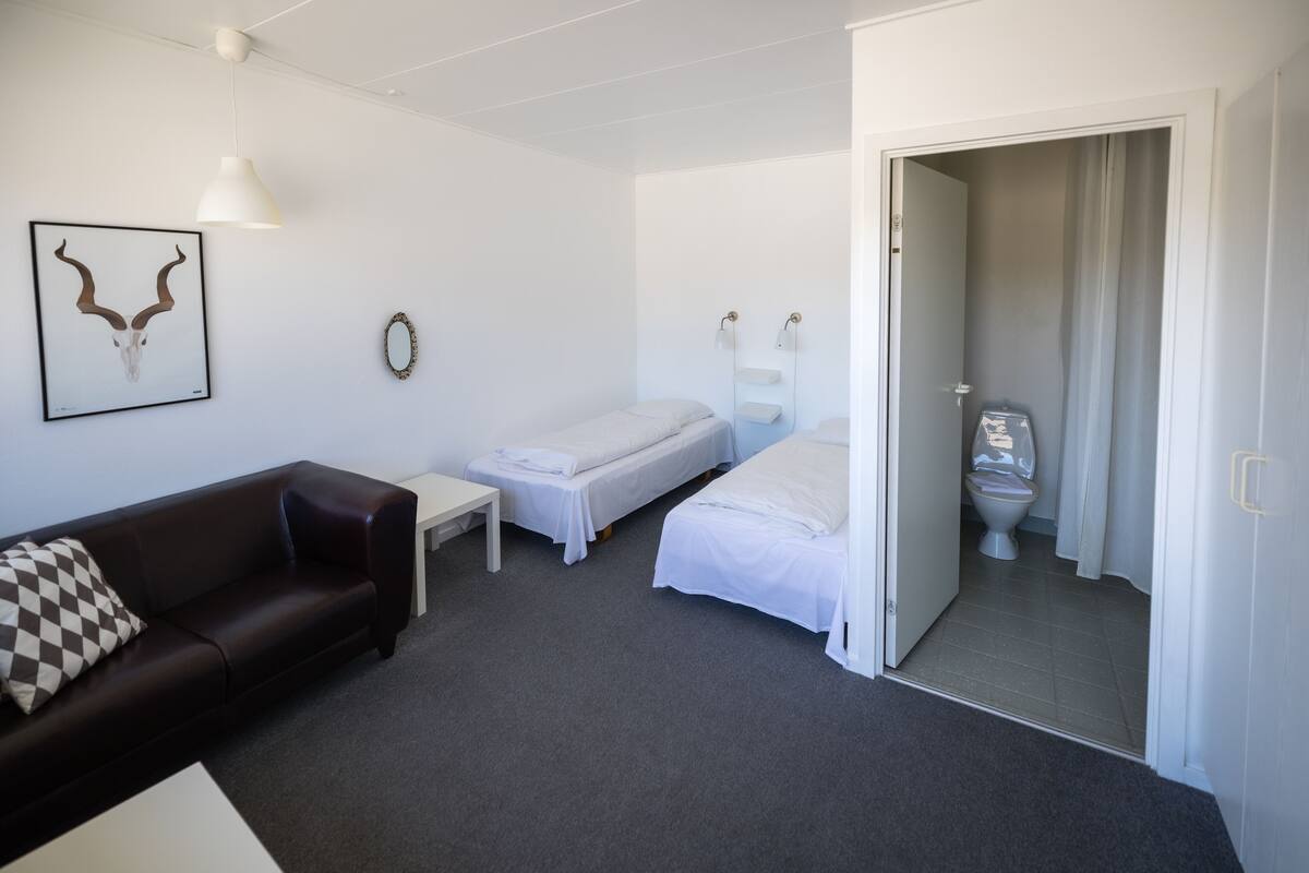 Une pièce avec un canapé en cuir marron, deux lits simples, une petite table, un tableau encadré au mur et une salle de bain visible à travers une porte ouverte.