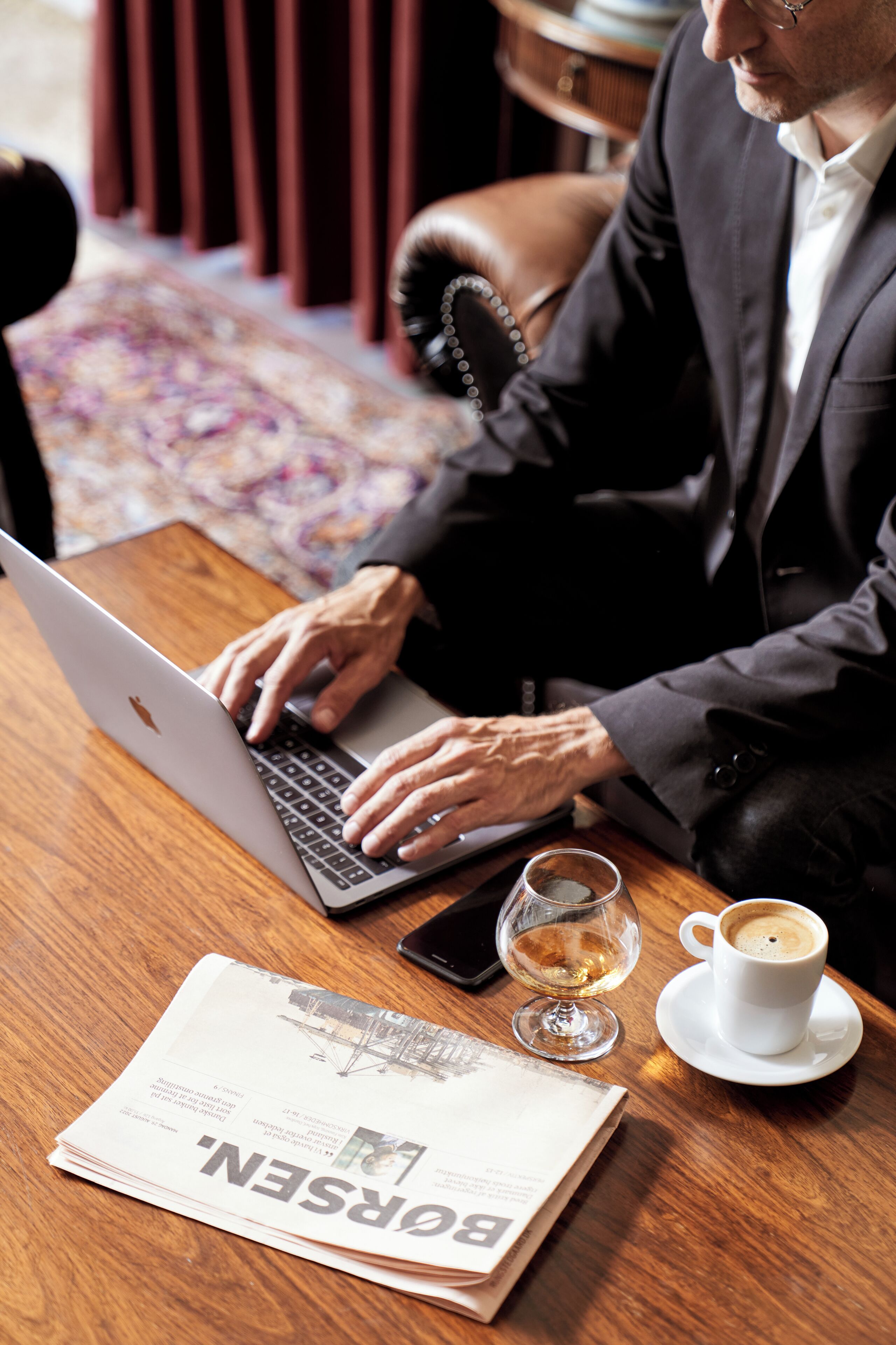 A person using a laptop at a wooden table with a newspaper, a glass of wine, and a cup of coffee.