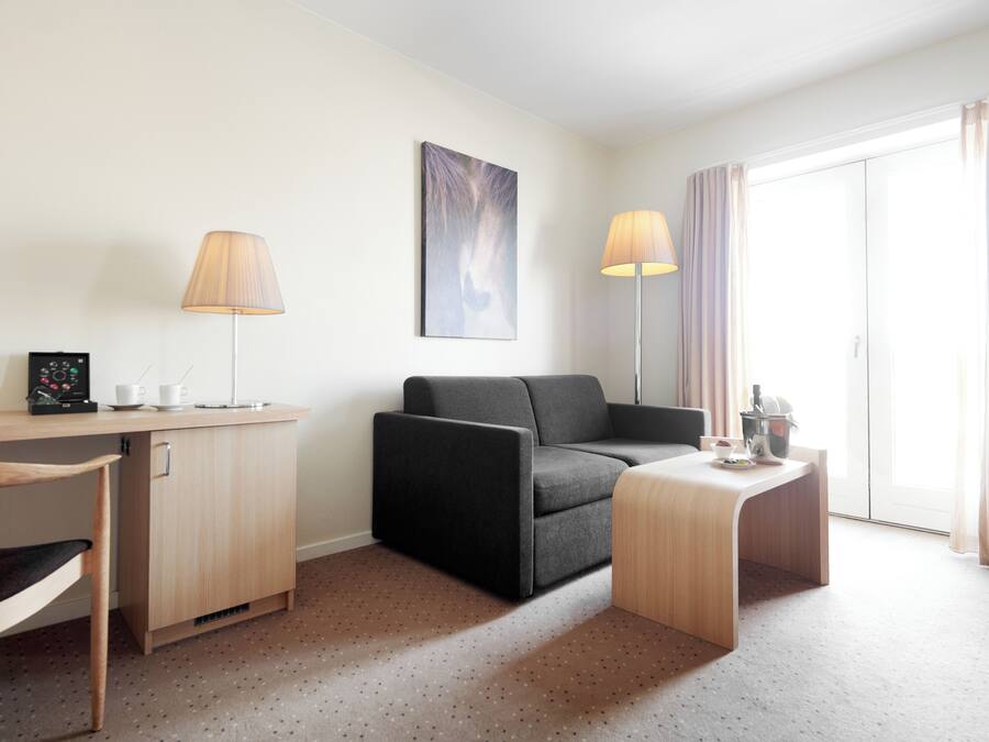 A modern hotel room with a wooden desk, a black sofa, a small wooden table, and a large window with curtains.