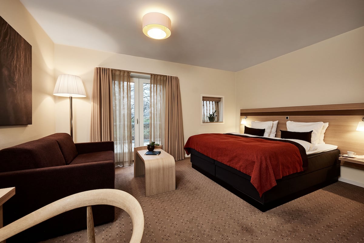 A hotel room with a bed, a sofa, a small table, a lamp, and a window with curtains.