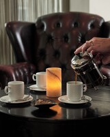 Una persona sirviendo café en una taza sobre una mesa con una vela, una silla y un plato de galletas.