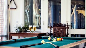 A pool table with balls and a pool cue, a wooden chair, and a table with glasses of wine and a plant.