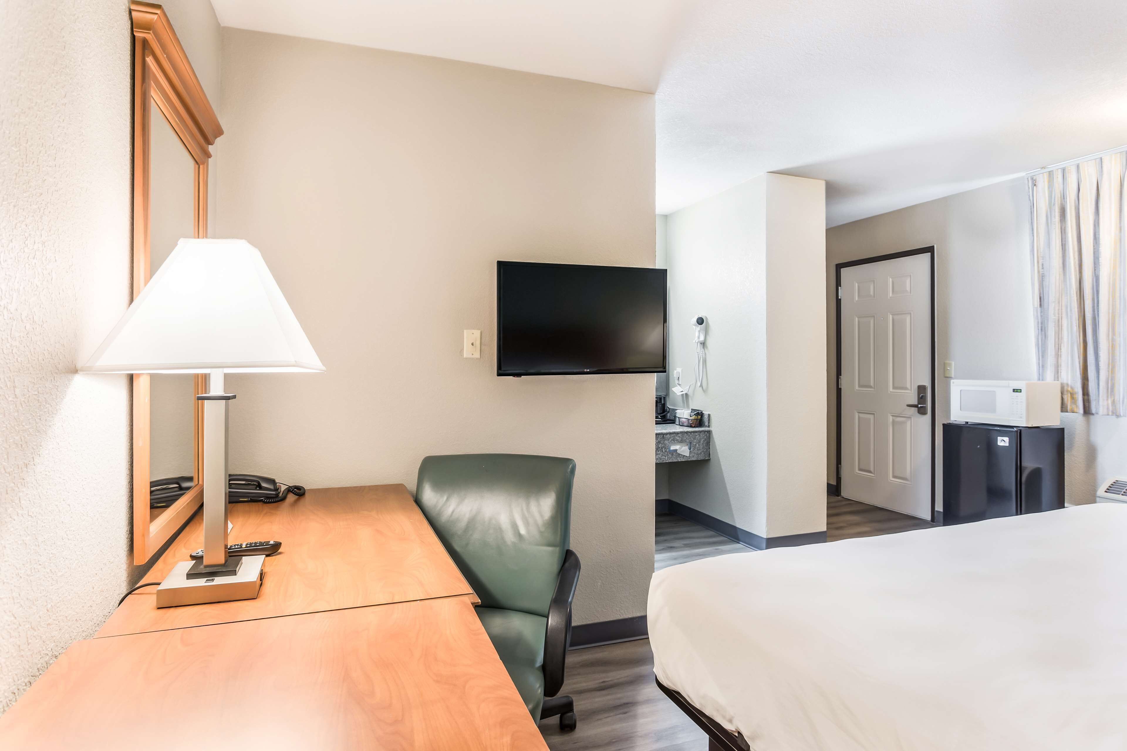 Habitación de hotel con cama, escritorio con lámpara, silla verde, televisión montada en la pared y una pequeña cocina equipada con microondas y refrigerador.
