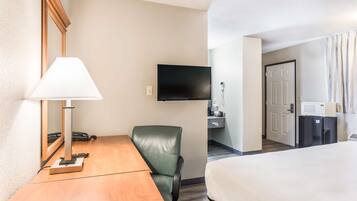 Habitación de hotel con cama, escritorio con lámpara, silla verde, televisión montada en la pared y una pequeña cocina equipada con microondas y refrigerador.