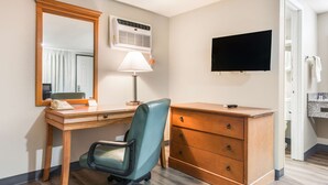 A hotel room with a wooden desk, a chair, a dresser, a mirror, a lamp, an air conditioner, and a TV.