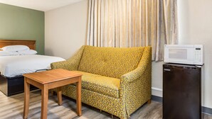 A hotel room with a bed, a yellow patterned armchair, a small wooden table, and a built-in microwave.