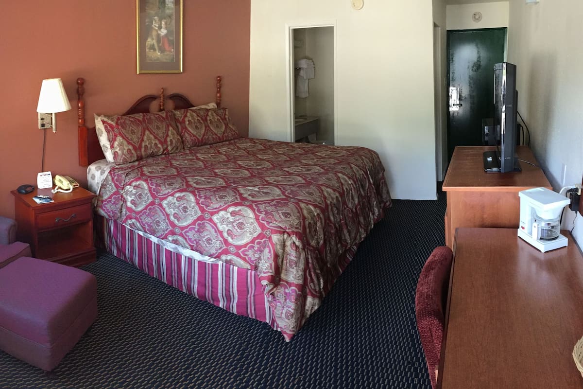 Hotellihuone, jossa on sänky, työpöytä, tuoli, yöpöytä lampun kanssa, puhelin ja lämmitin.
