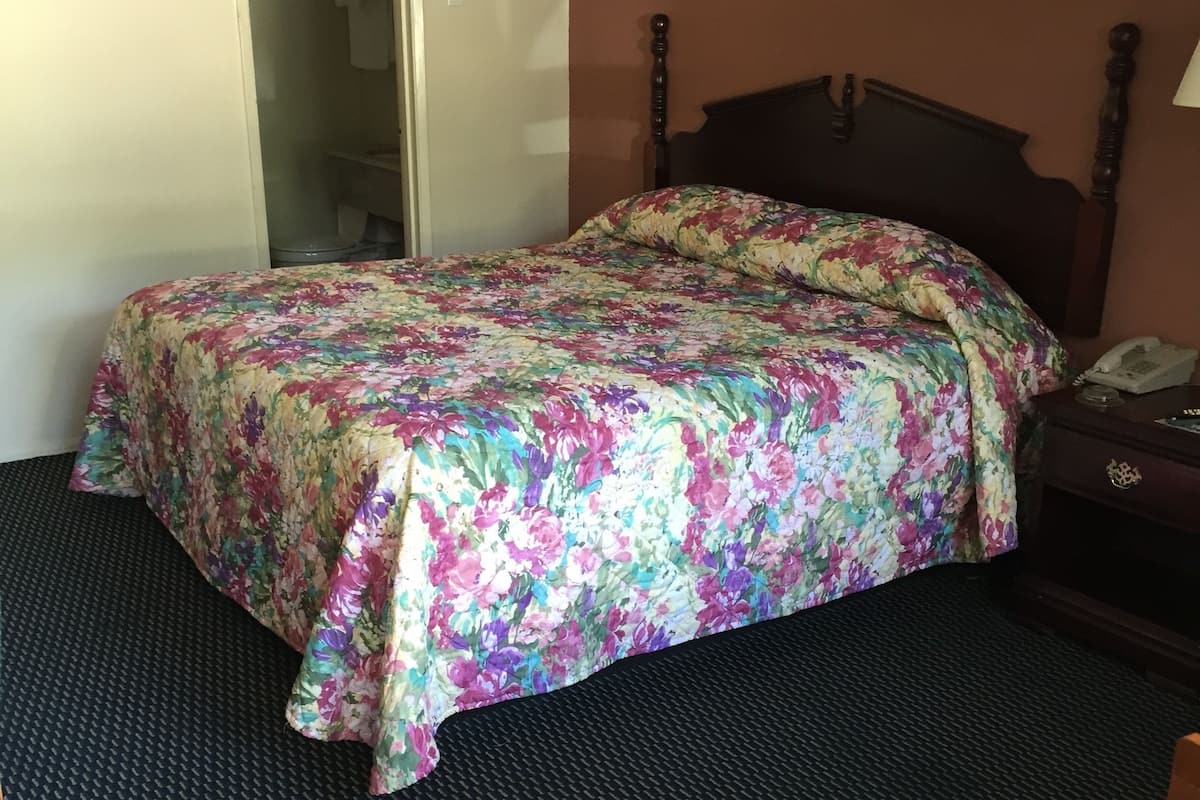 Hotellihuone, jossa on sänky, tuoli, yöpöytä, lamppu ja taulu seinällä.