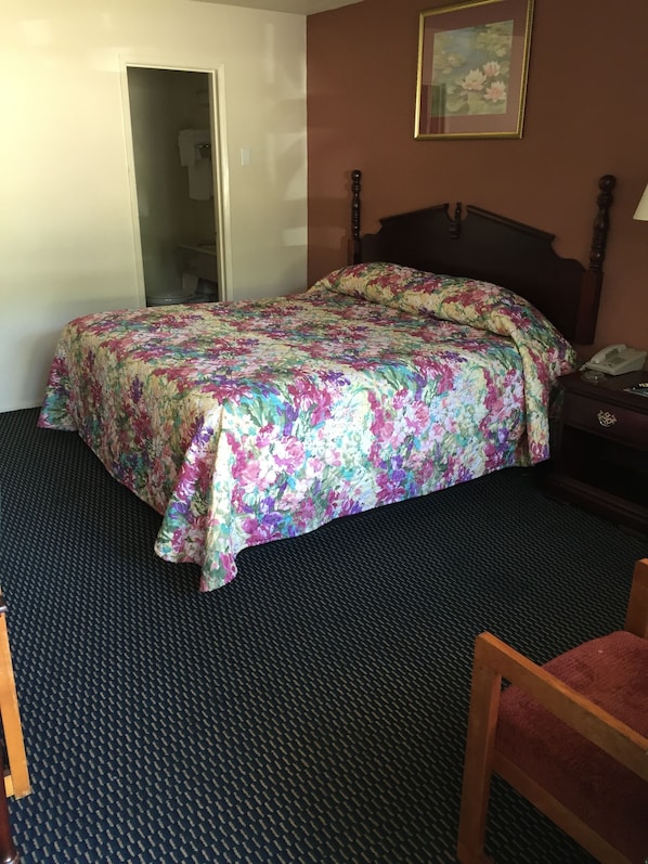 A hotel room with a bed, a chair, a nightstand, a lamp, and a painting on the wall. - Scottish Inns (Tupelo)