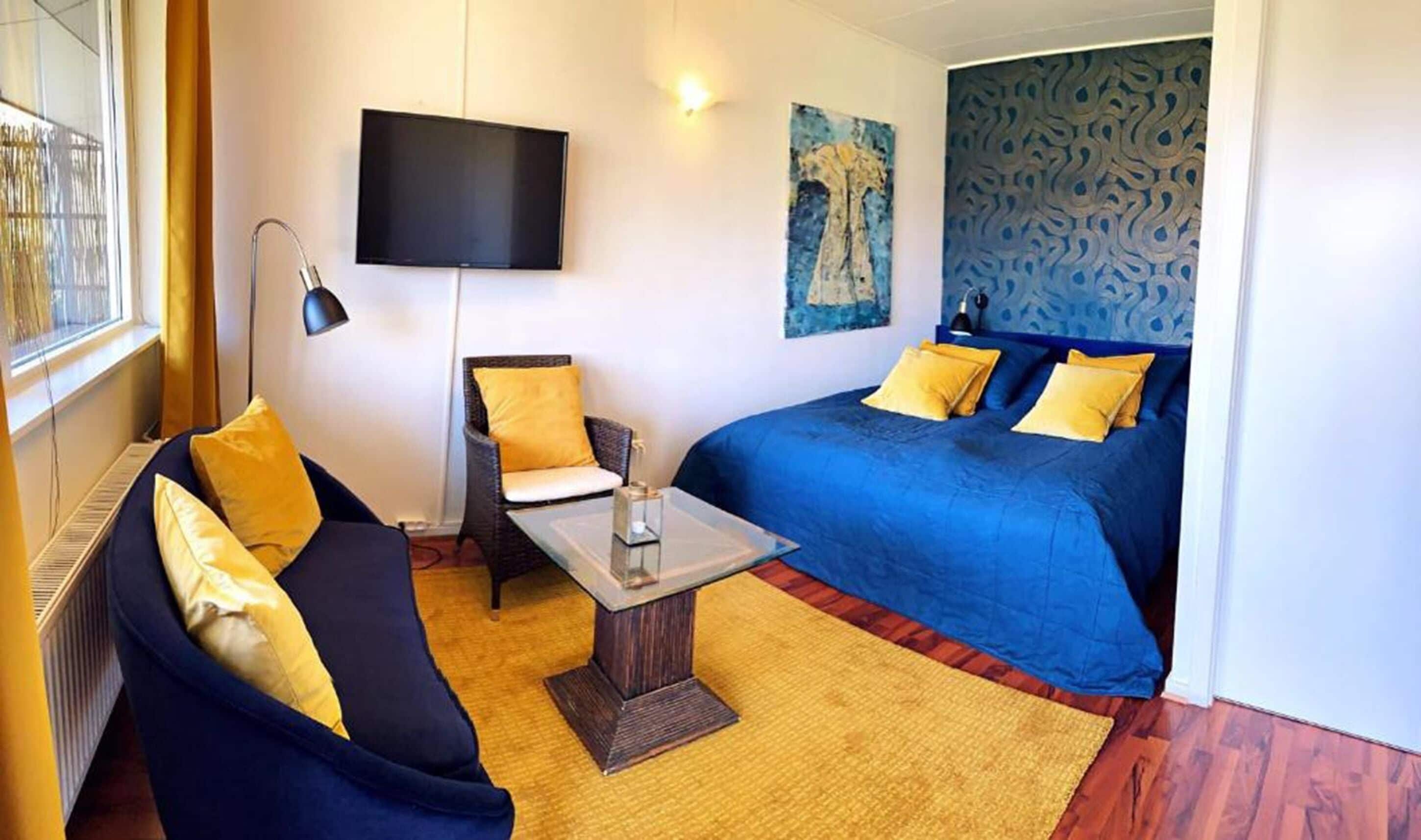 Habitación de hotel con sofá, cama con ropa de cama azul, mesita de noche con lámpara, televisor en la pared y un cuadro.