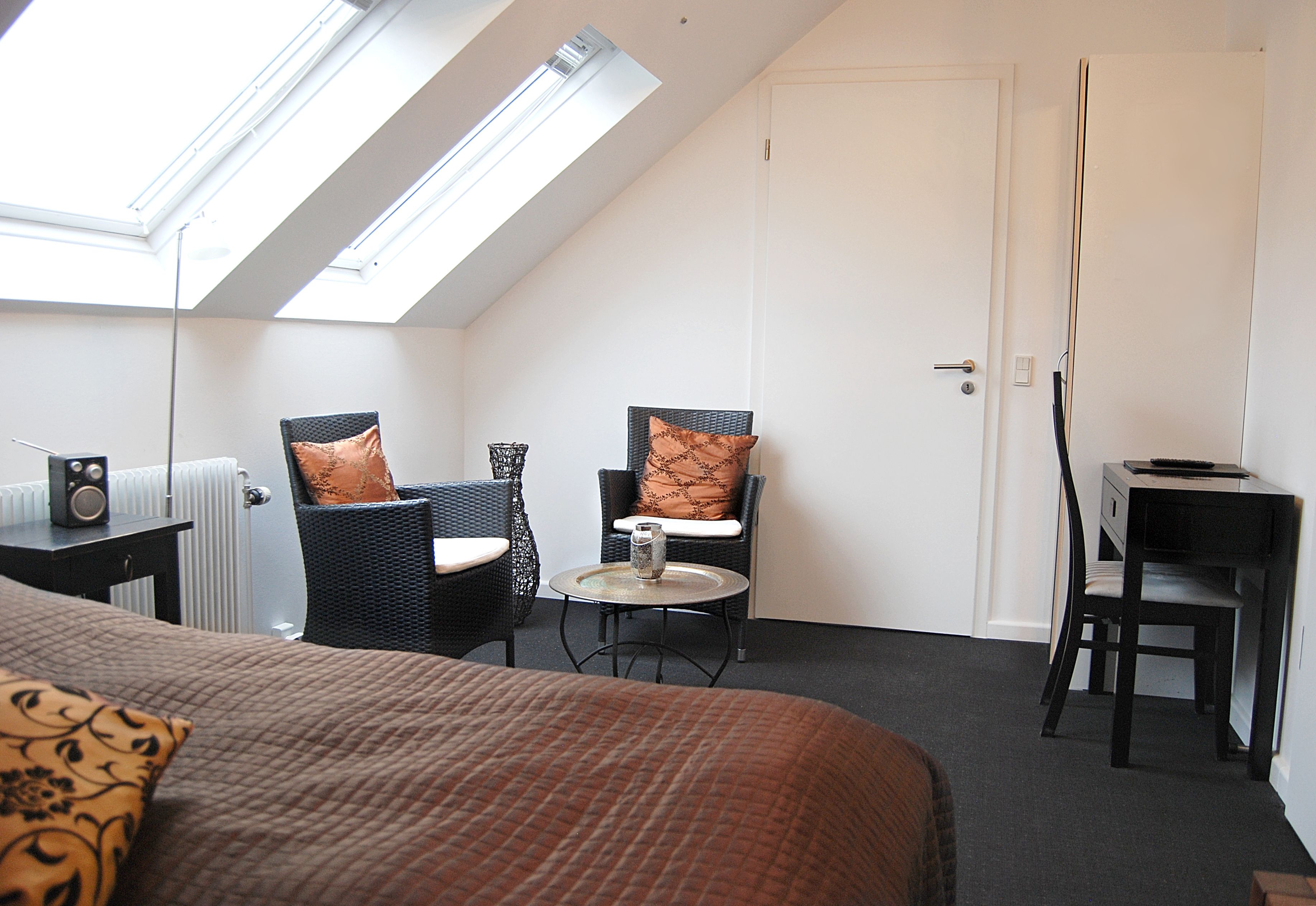 Une pièce comprenant un lit, deux chaises, une petite table et un bureau. Il y a une lucarne au-dessus du lit et une porte sur la droite.