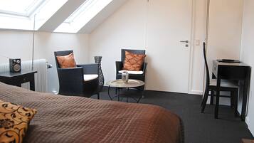 Une pièce comprenant un lit, deux chaises, une petite table et un bureau. Il y a une lucarne au-dessus du lit et une porte sur la droite.