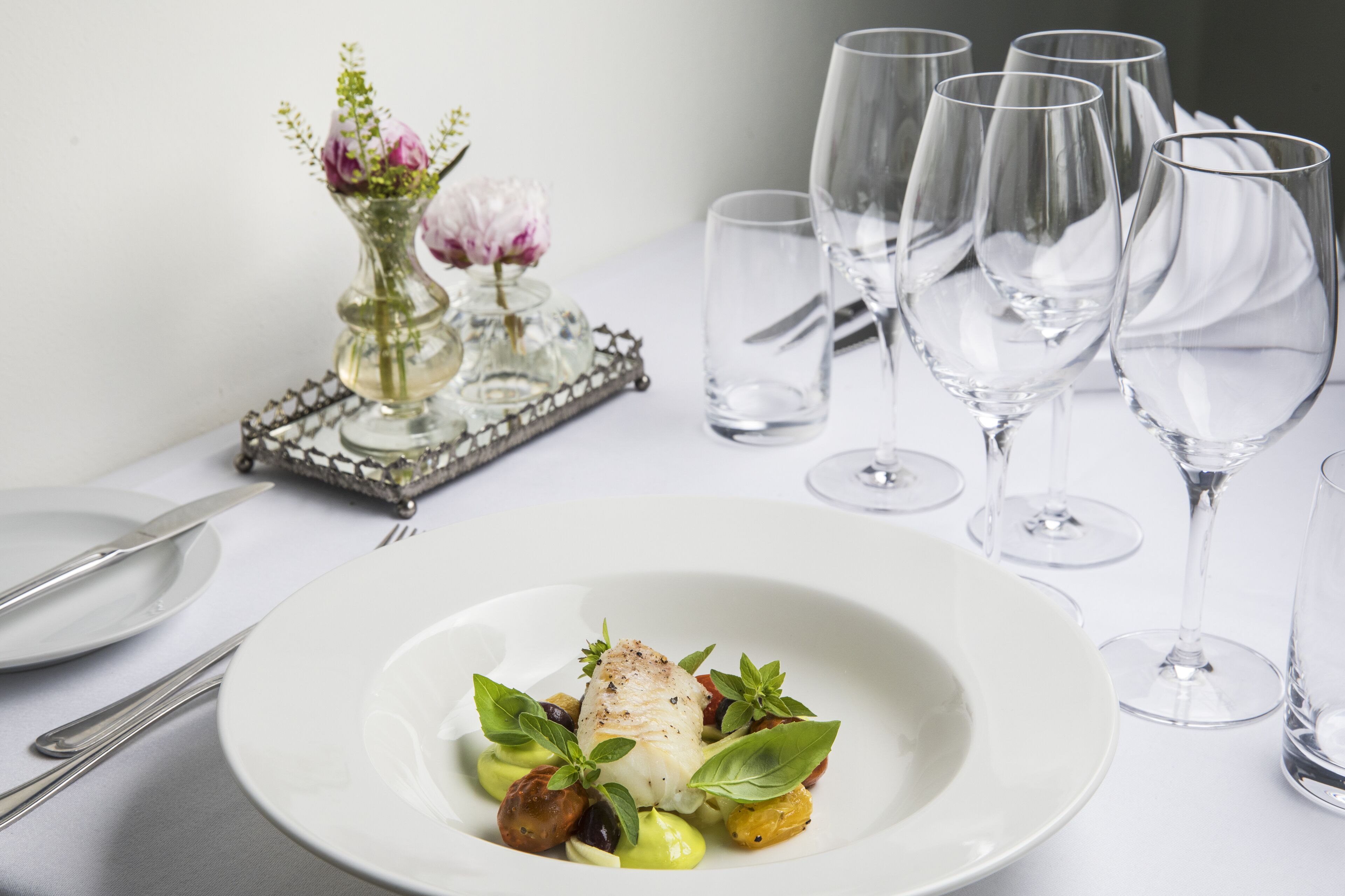 Una mesa de comedor dispuesta con un plato blanco de comida, copas de vino transparentes y un florero decorativo con flores.