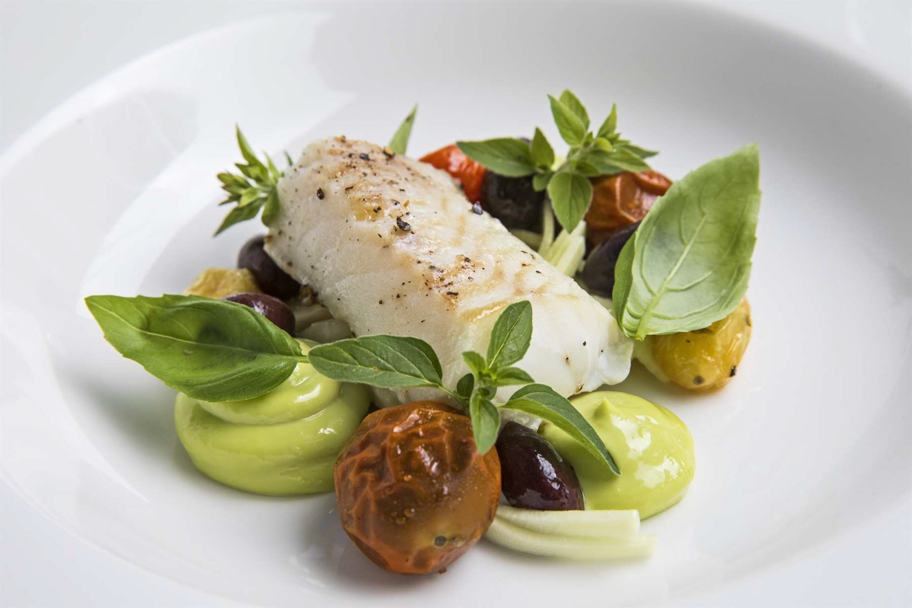 A white plate with a piece of white fish, green sauce, olives, and herbs.